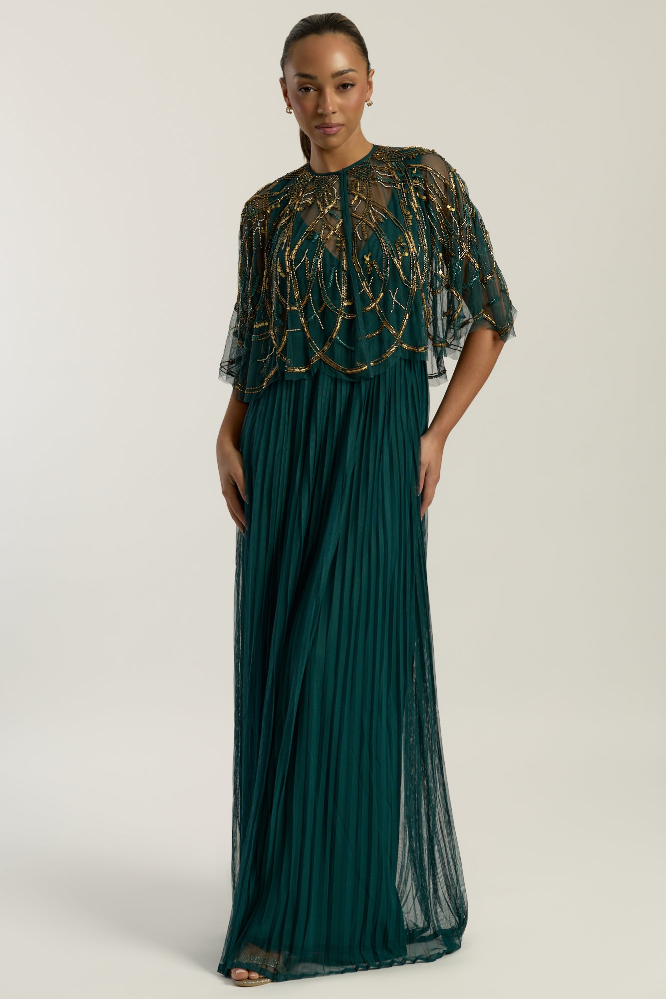 Green embellished maxi dress with flowing pleated skirt and detachable sheer cape overlay, featuring gold bead detailing and an elegant V-neck silhouette for occasion wear.