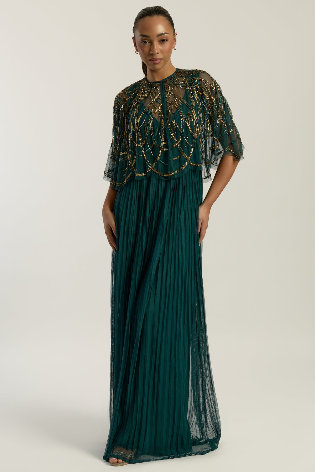 Green embellished maxi dress with flowing pleated skirt and detachable sheer cape overlay, featuring gold bead detailing and an elegant V-neck silhouette for occasion wear.
