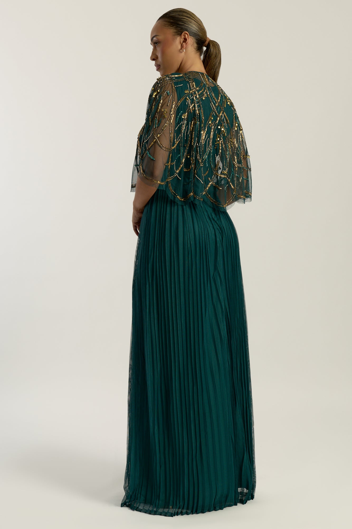 Green embellished maxi dress with flowing pleated skirt and detachable sheer cape overlay, featuring gold bead detailing and an elegant V-neck silhouette for occasion wear.